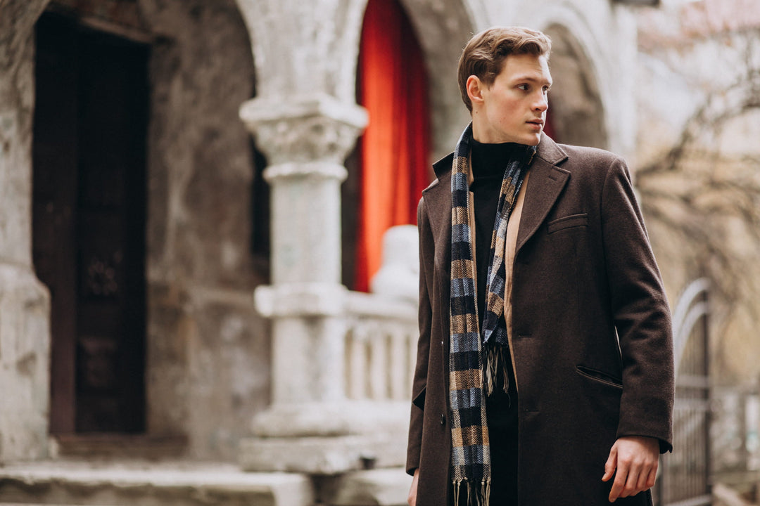 A young man wearing a classic dark brown wool overcoat and a checkered heritage scarf, standing in front of traditional stone architecture with arched pillars.